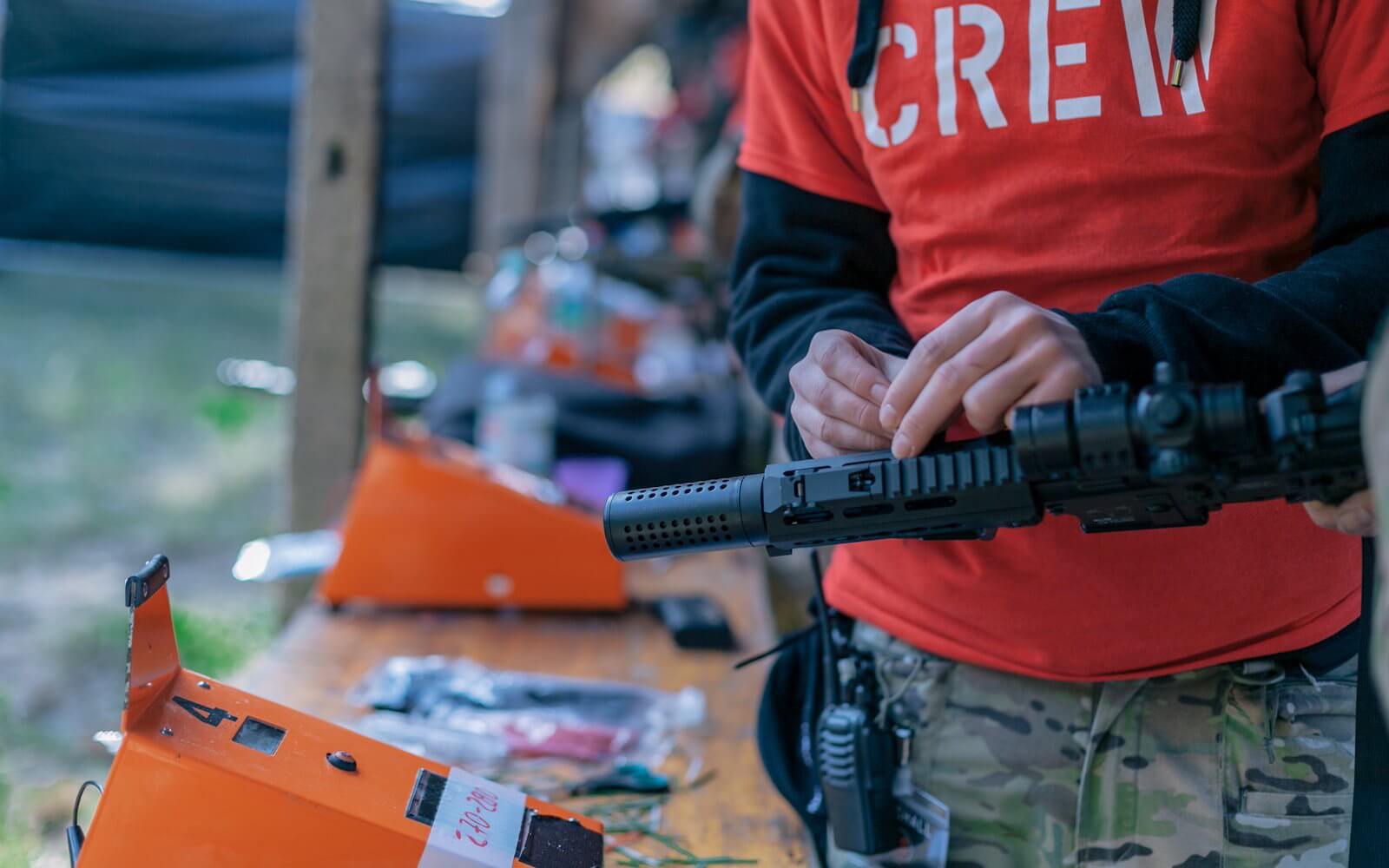 Person justiert Airsoft-Waffe in rotem "CREW"-Shirt, vor orangefarbenen Geräten.
