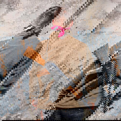 Person mit Airsoft-Gewehr auf dem Rücken, trägt braune Jacke und rotes Halstuch, steht vor verputzter Wand.