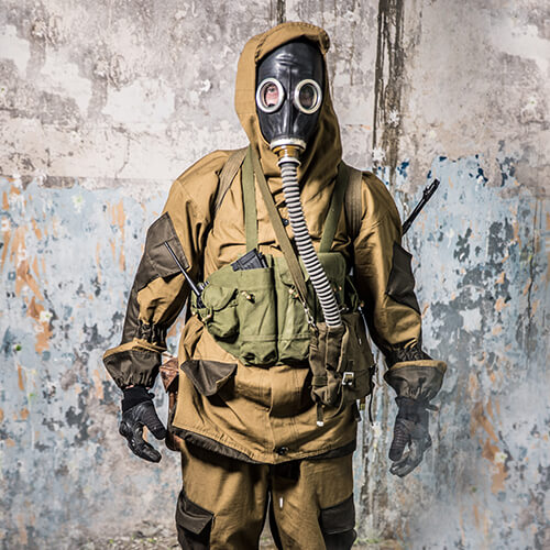 Person in Tarnanzug mit Gasmaske und taktischem Equipment vor einer zerfallenen Wand.