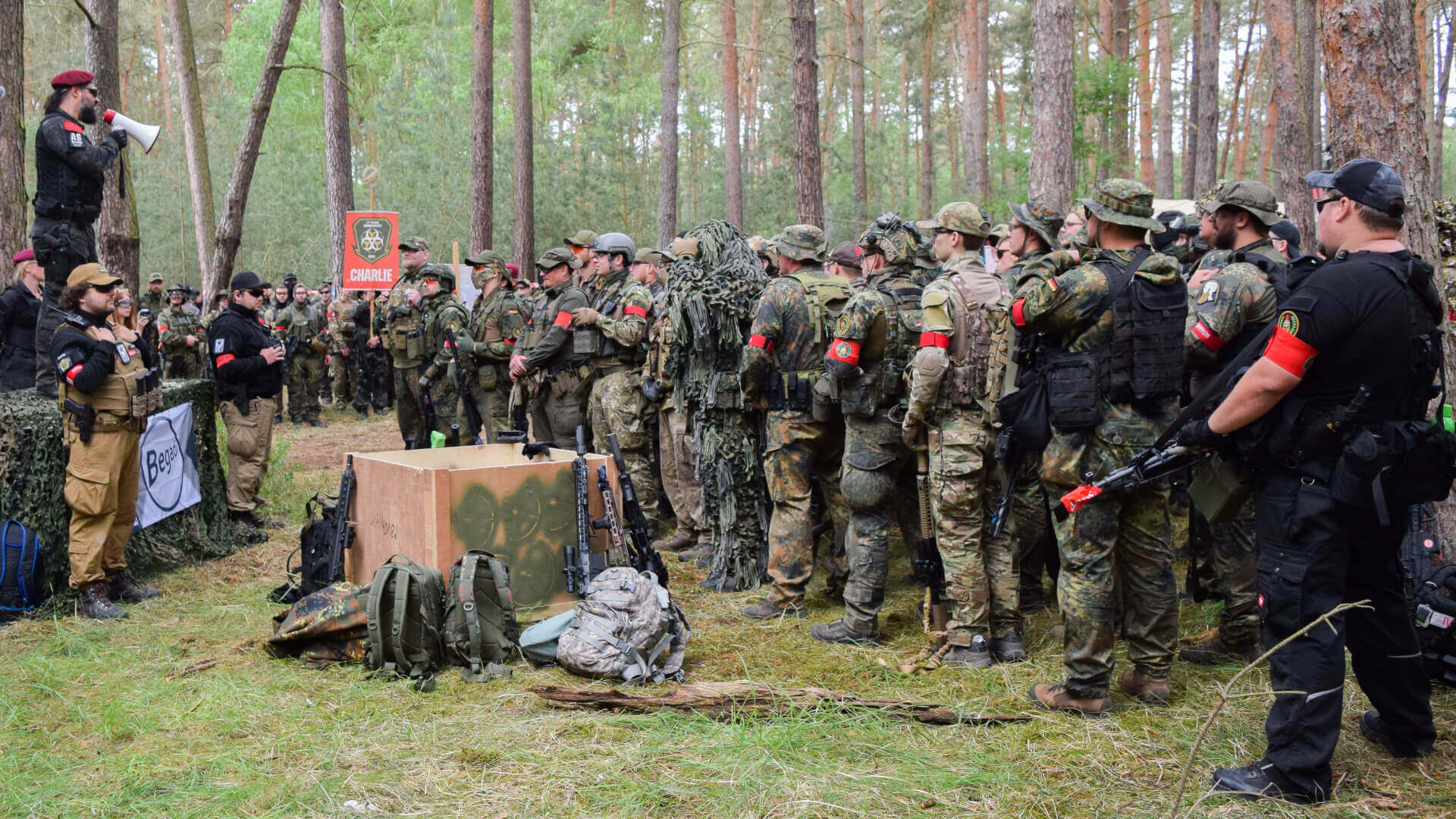 Airsoft-Teambriefing im Wald: Spieler in Tarnkleidung mit Markierern, ein Anführer spricht per Megafon.