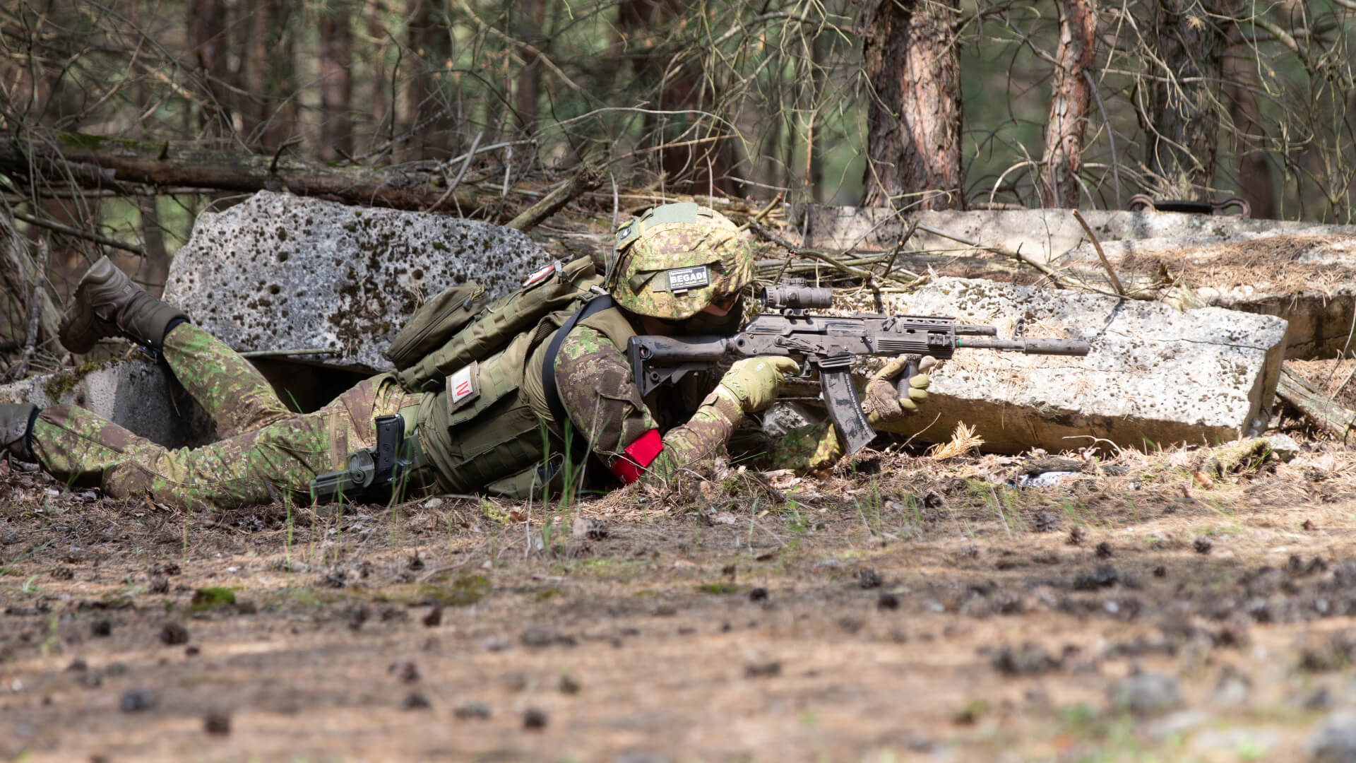 Airsoft-Spieler in Tarnkleidung zielt liegend mit Gewehr im Wald.