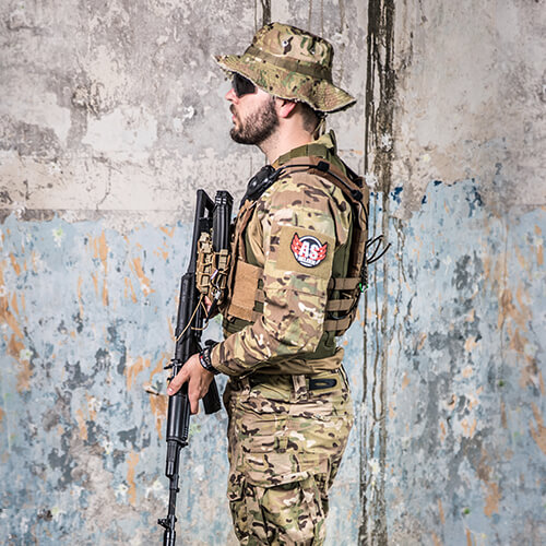 Airsoft-Spieler in Tarnkleidung mit Gewehr vor einer verwitterten Wand.