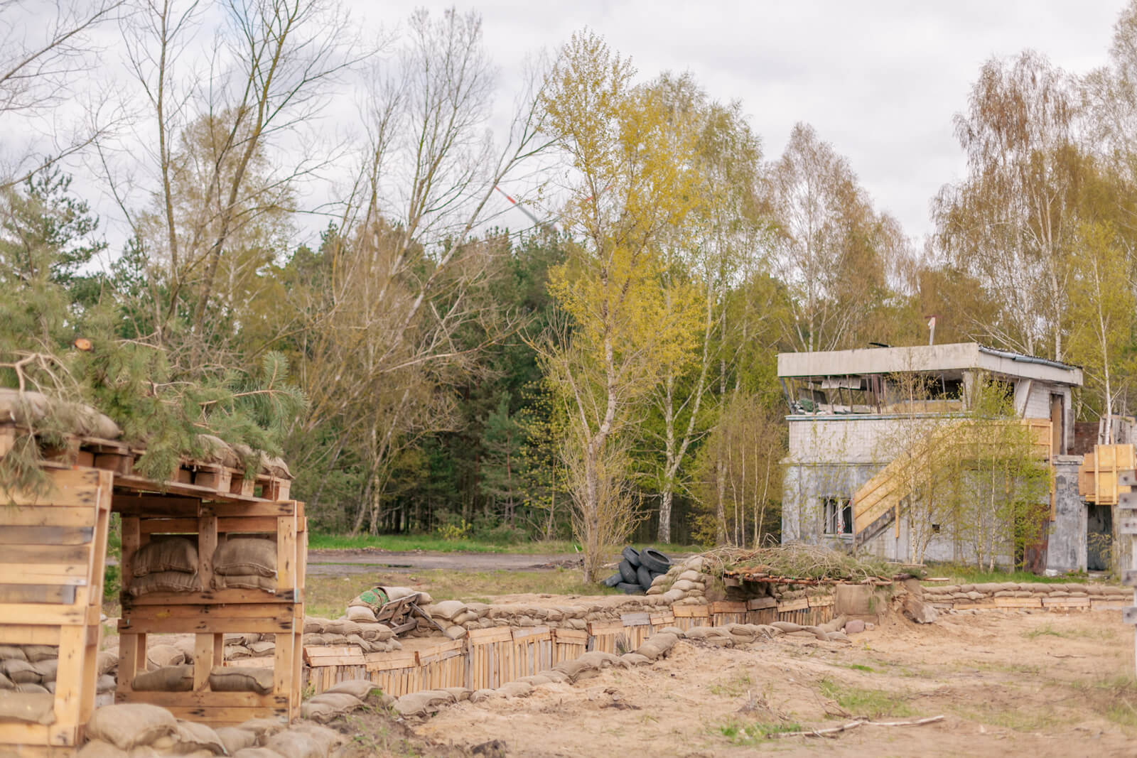 Airsoftfeld mit Schützengraben, Sandsäcken und Gebäude vor herbstlichem Wald.