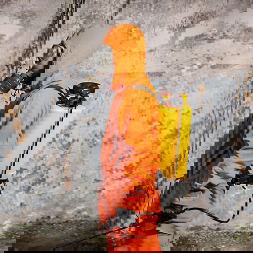 Person in orangem Schutzanzug mit Gasmaske und Sprühgerätrucksack steht vor einer verwitterten Wand.