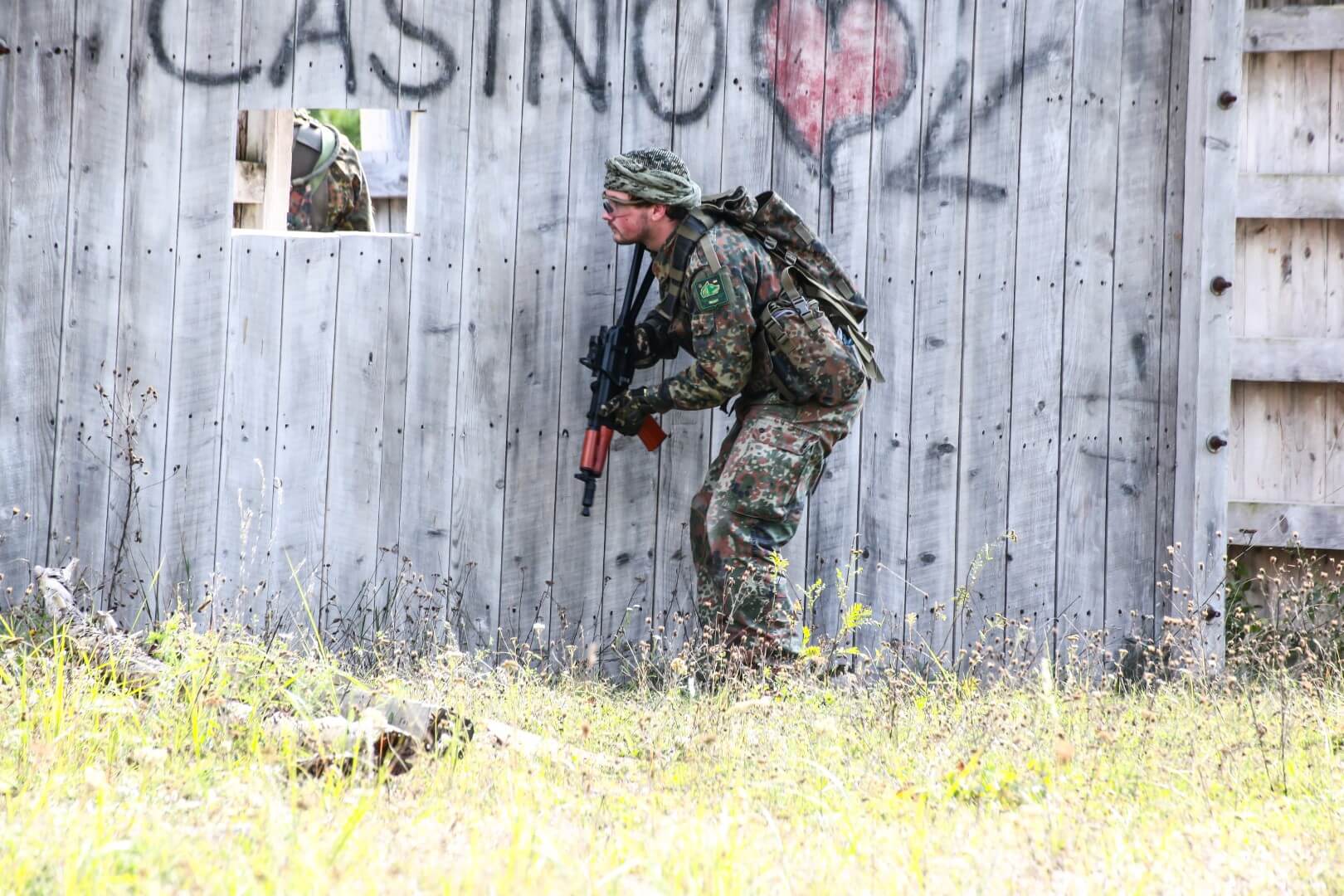 Person in Tarnuniform hält Airsoft-Gewehr, kauert neben Holzwand mit Graffiti.