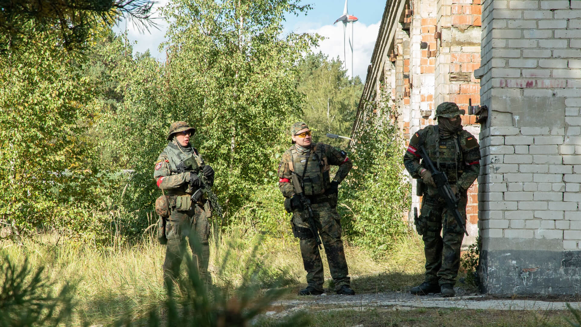 Drei Airsoft-Spieler in Tarnkleidung neben einer Ruine im Wald. Einer lehnt an der Wand mit Gewehr.