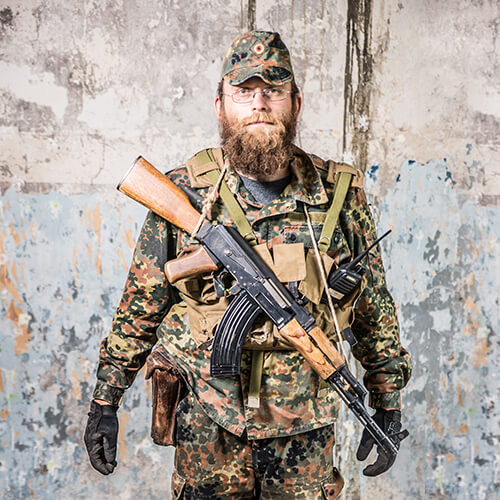 Person in Tarnkleidung mit Airsoft-Gewehr und Funkausrüstung vor rustikalem Hintergrund.