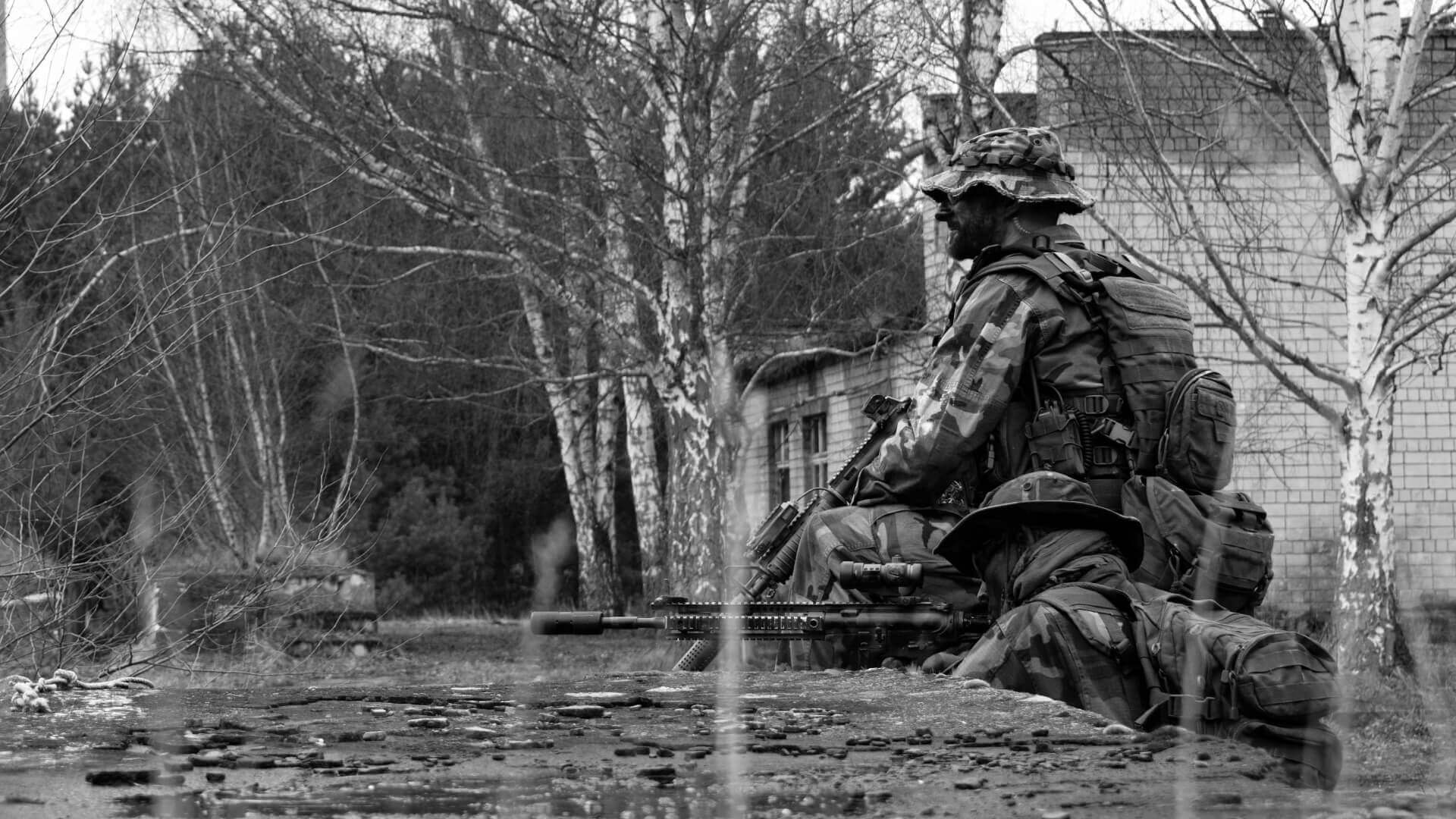 Zwei Airsoft-Spieler in Tarnkleidung zielen hinter Deckung in einer bewaldeten, urbanen Umgebung.