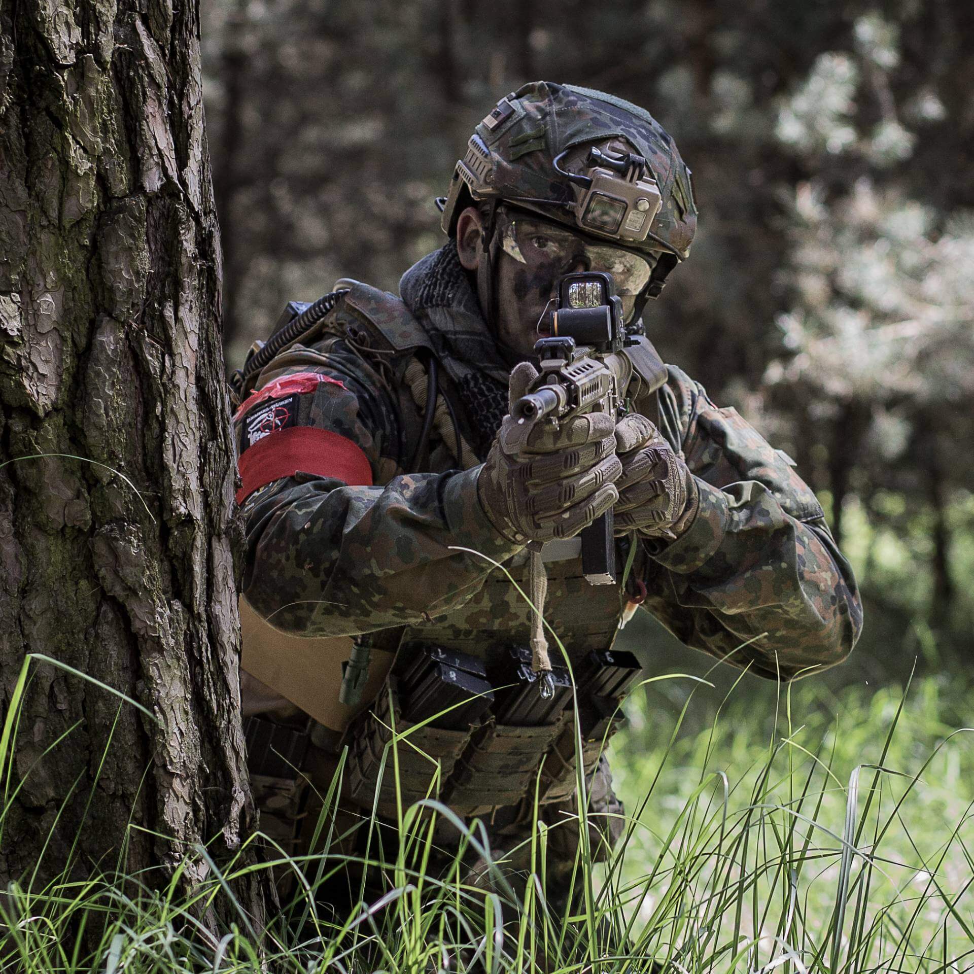 Airsoft-Spieler in Tarnkleidung zielt im Wald hinter einem Baum hervor.
