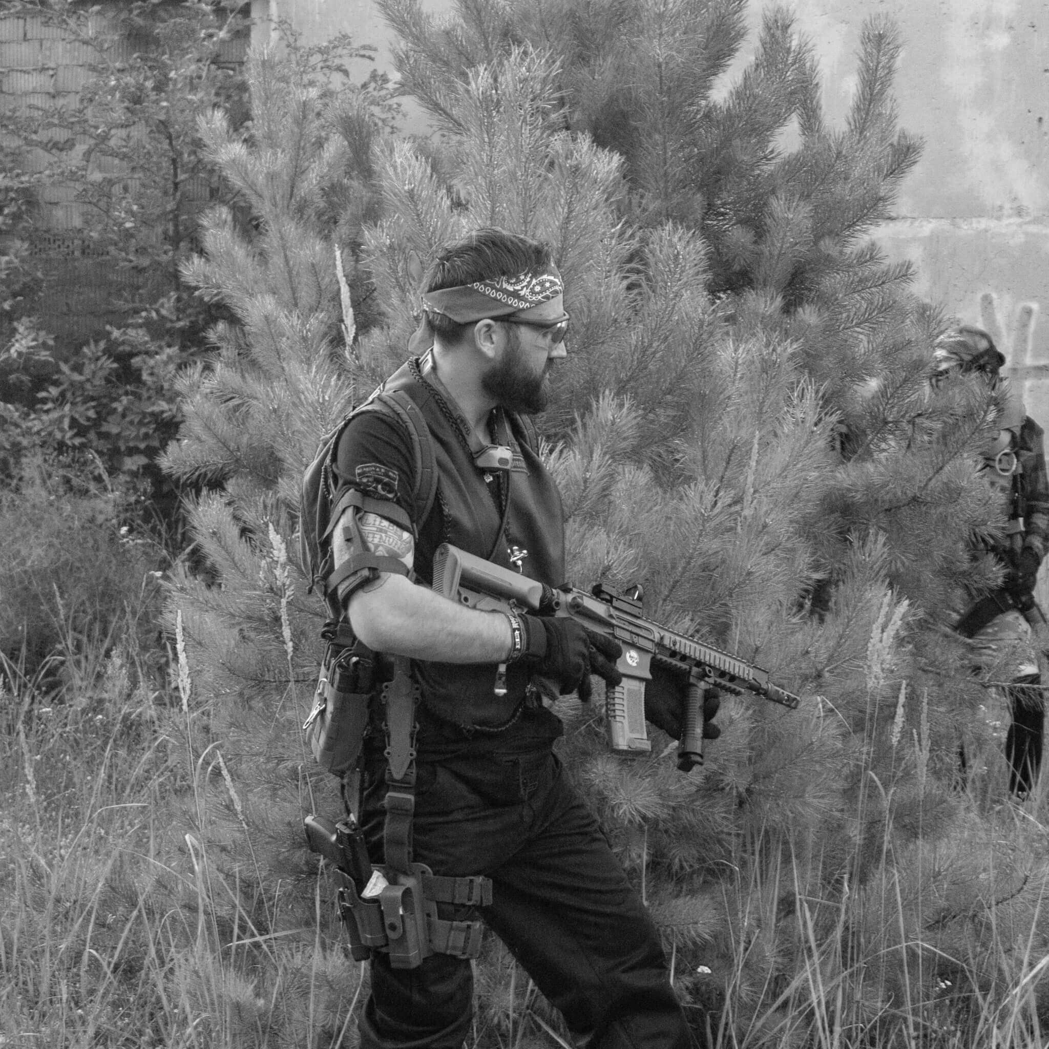 Person mit Airsoft-Gewehr und taktischer Ausrüstung in bewaldetem Gelände. Schwarz-Weiß-Foto.