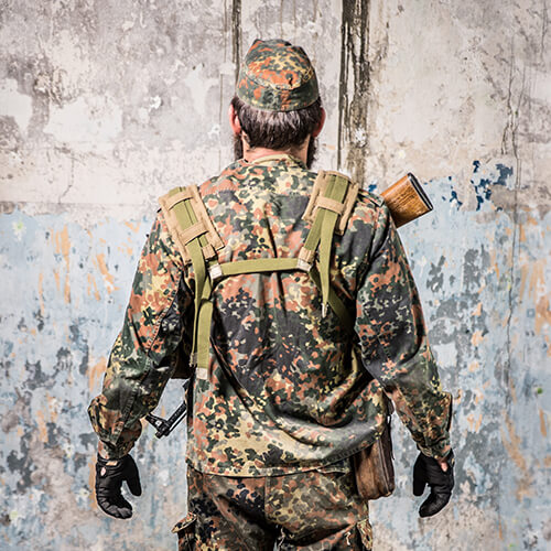 Person in Flecktarn-Uniform mit Gewehr, stehend vor einer verwitterten Wand; Rückansicht.
