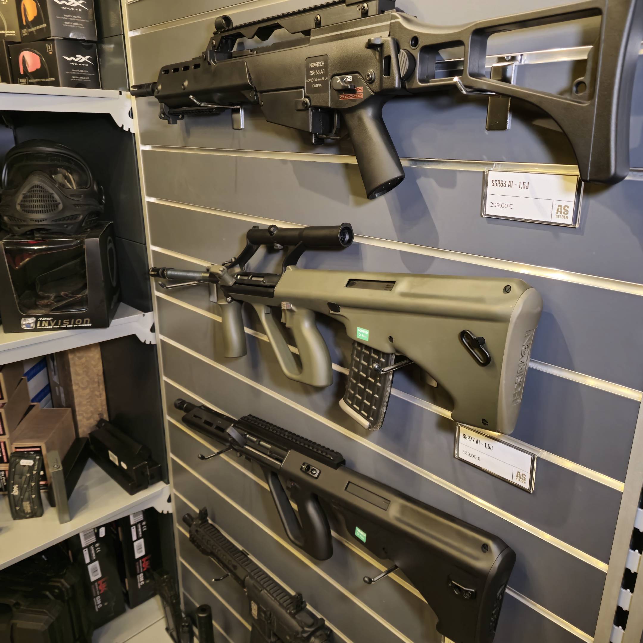 Airsoft-Waffen an einer Wand ausgestellt, darunter ein Modell mit Zubehör und Preisschilder.