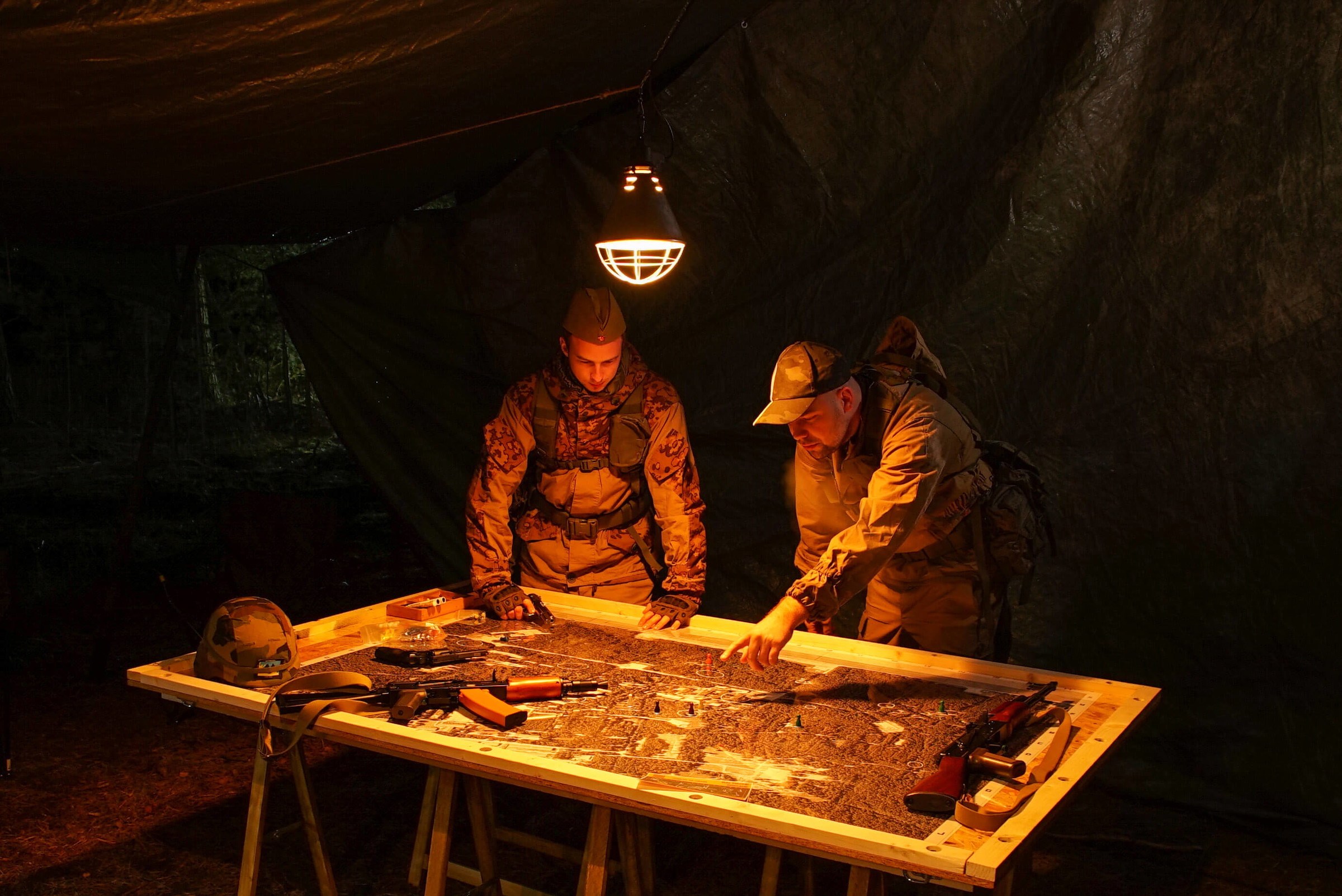Zwei Airsoft-Spieler planen unter einer Lampe eine Strategie an einem Tisch mit Karten und Ausrüstung.