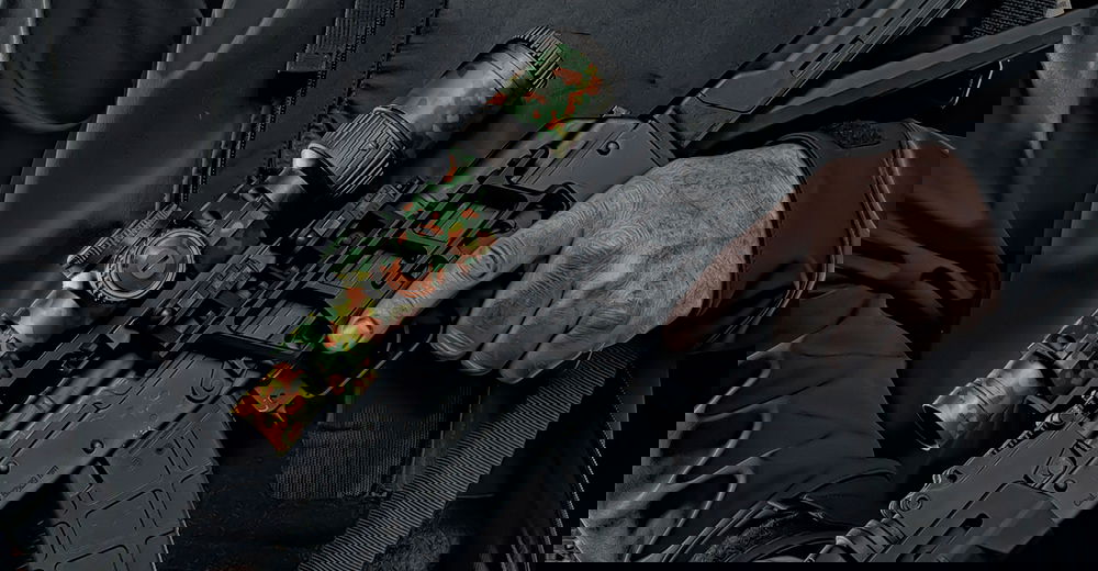 GunsWrap Scope Skins Person hält Airsoft-Gewehr mit Camouflage-Zielfernrohr, schwarze Kleidung.