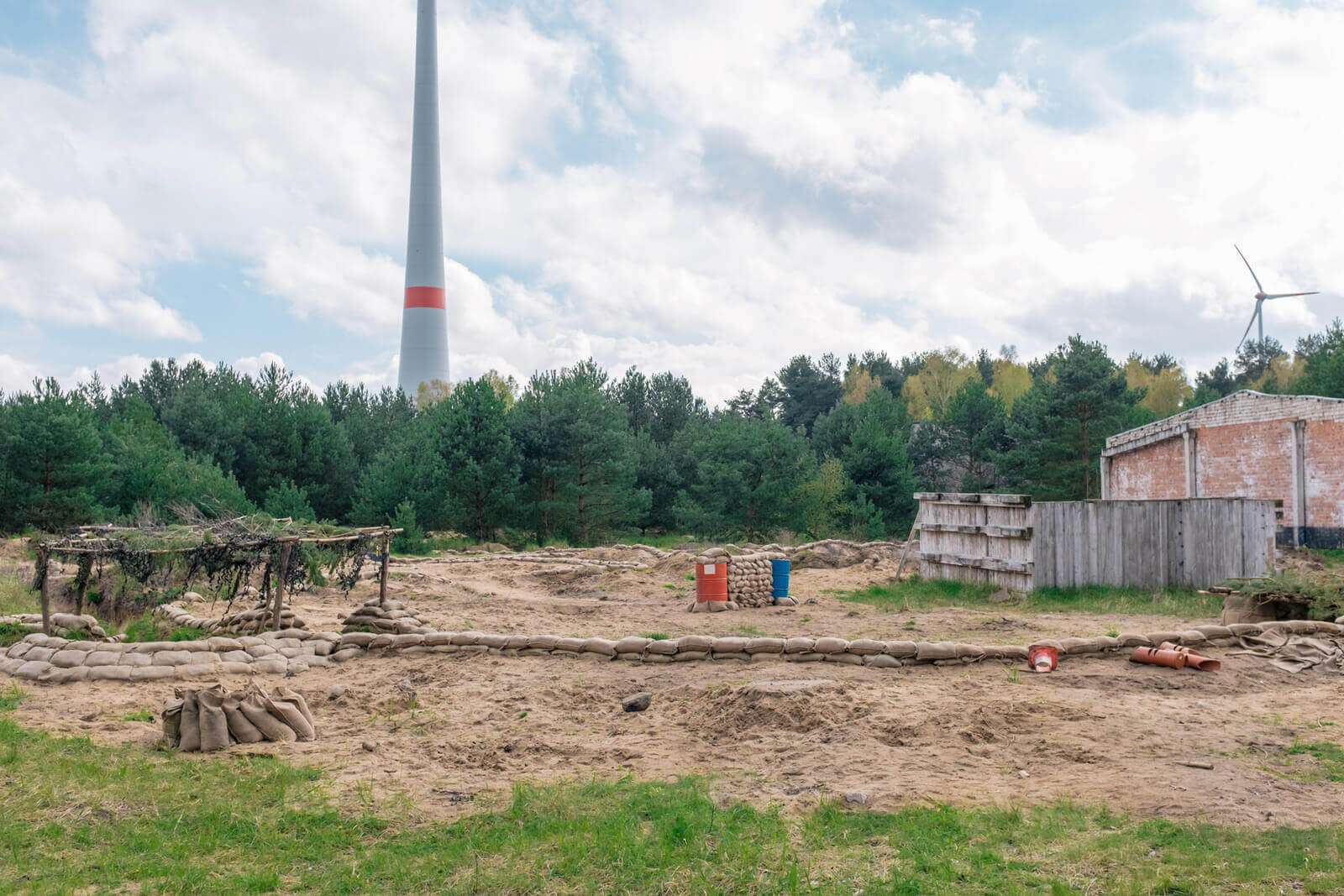 Airsoft-Feld mit Sandsackdeckungen, windige Landschaft, Bunker und Bäume im Hintergrund.