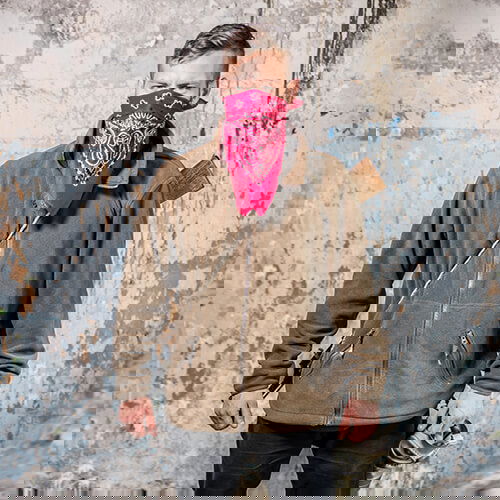 Person mit rotem Tuch und Airsoft-Ausrüstung vor rustikalem Hintergrund.